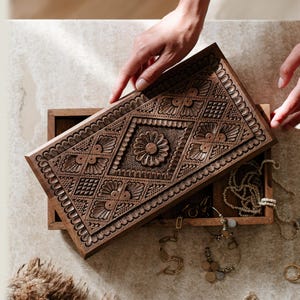 May include: A rectangular wooden jewelry box with a hinged lid featuring intricate hand-carved floral and geometric patterns. The dark brown wood lid is partially open, revealing an interior compartment containing various gold-toned necklaces and rings.