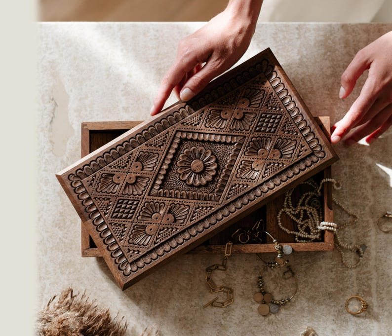 May include: A rectangular wooden jewelry box with a hinged lid featuring intricate hand-carved floral and geometric patterns. The dark brown wood lid is partially open, revealing an interior compartment containing various gold-toned necklaces and rings.