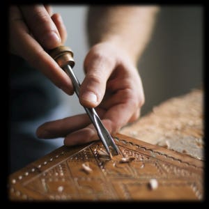 May include: A metal gouge tool with a wooden handle is being used to carve intricate geometric patterns into a flat, tan-colored leather surface. Small shavings of leather are visible on the surface as the tool creates a decorative design.