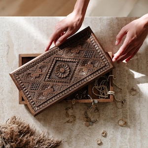 May include: A detailed wooden jewellery box with a carved lid. The lid is being opened to reveal jewellery, including necklaces and rings. The box is brown and rests on a light-coloured surface.