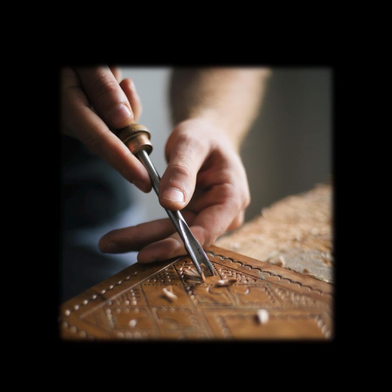 Puede incluir: Una herramienta de metal para trabajar el cuero con mango de madera que se utiliza para tallar patrones geom&eacute;tricos intrincados en una superficie de cuero color canela, creando peque&ntilde;as virutas a medida que la punta de metal corta el material.