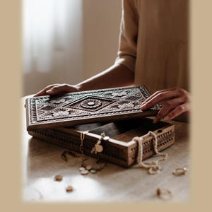 Puede incluir: Un joyero rectangular de madera marr&oacute;n oscura con una tapa intrincadamente tallada que presenta un motivo floral central y patrones geom&eacute;tricos. La caja est&aacute; abierta, revelando un compartimento interior vac&iacute;o, con cadenas doradas y anillos cerca.