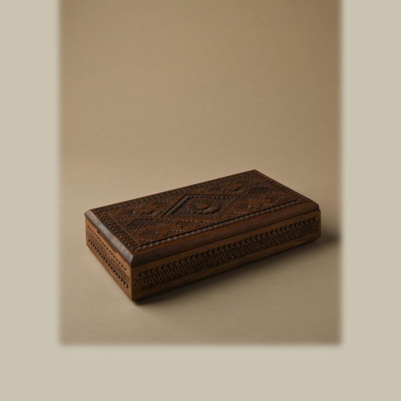 Puede incluir: Una caja rectangular de madera con intrincados patrones geom&eacute;tricos tallados a mano y un motivo floral central en la tapa. La superficie de madera marr&oacute;n oscuro muestra grabados detallados a lo largo de la parte superior y los laterales.