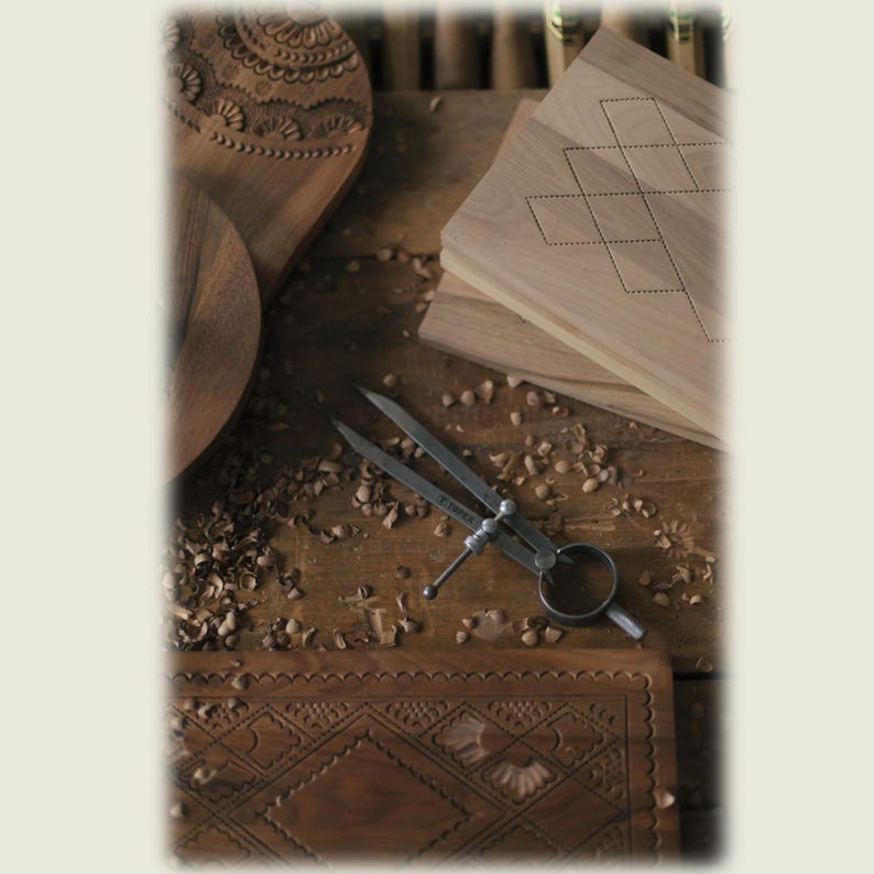 May include: A metal divider tool with a ring handle and two pointed legs rests on a wooden surface alongside several light and dark brown wooden boards. The boards feature intricate carved geometric patterns and dotted line designs.