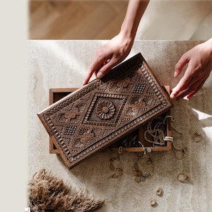 Puede incluir: Un joyero rectangular de madera con un intrincado patr&oacute;n floral y geom&eacute;trico tallado a mano en la tapa. La tapa est&aacute; parcialmente abierta, revelando un compartimento de almacenamiento que contiene varios collares, anillos y joyas doradas.