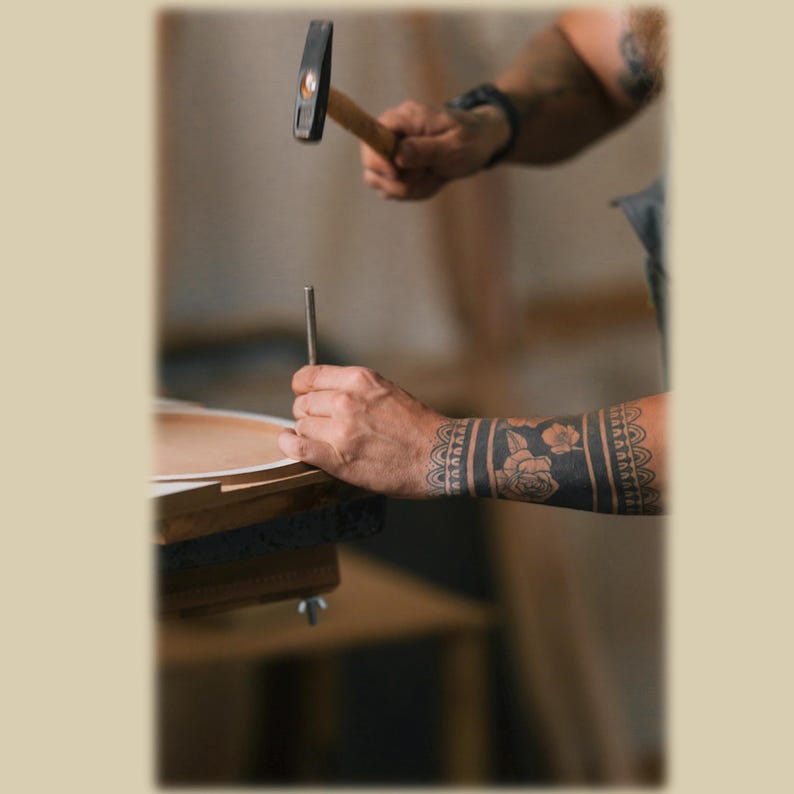 May include: A metal hammer held above a thin metal rod positioned on a circular wooden surface. The arm holding the hammer features a dark floral tattoo band around the forearm, while the other hand stabilizes the rod against the wood.