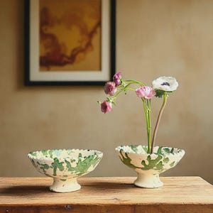 Op de afbeelding: Twee keramische voetstukkommen met een crèmekleurige basis en een groen spetterontwerp. Eén kom bevat roze en witte bloemen. De kommen staan op een houten tafel. Een ingelijst schilderij hangt op de achtergrond.