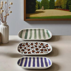 May include: Three oval serving platters with different patterns. One has green stripes, another has a leopard print, and the third has purple stripes. The platters are white with a raised edge.