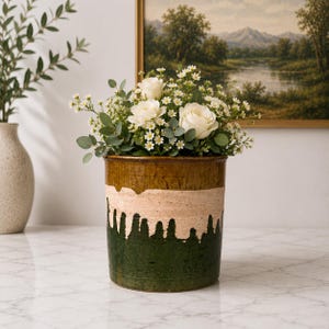 May include: A cylindrical ceramic vase featuring a tri-color glaze with a dark brown top, a central band of textured beige, and a deep green base with a drip pattern. The vase is filled with a bouquet of white roses, small white flowers, and eucalyptus.