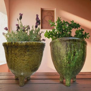 May include: Two green ceramic planters with three legs each, filled with plants. One planter holds lavender with purple flowers, the other has green leafy plants. The planters have a textured, aged appearance, sitting on a wooden surface.