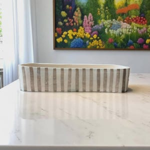 Könnte beinhalten: Ein rechteckiger Keramik-Blumentopf mit cremefarbenem Grund und vertikalen taupefarbenen Streifen. Der Topf steht auf einer weißen Marmoroberfläche, mit einem farbenfrohen Blumenbild im Hintergrund.