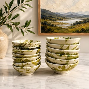 May include: Two stacks of ceramic bowls featuring an off-white base colour with irregular, hand-painted green splatters and streaks. Each bowl has a rounded shape with a small footed base, showcasing a consistent artistic pattern across all pieces.