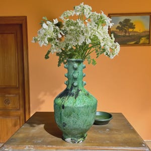 May include: A tall, bulbous green ceramic vase with a glossy, mottled glaze and a series of small, raised circular protrusions along the neck. The vase is filled with a large bouquet of white flowers with delicate, lacy green foliage.