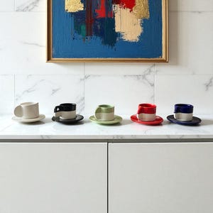 May include: Five ceramic coffee cups and saucers in a row. Each cup has a unique color scheme: cream, black and white, green and cream, red and cream, and blue and cream. The cups are on a white countertop with a colorful abstract painting in the background.