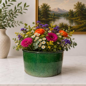 Puede incluir: Un vibrante arreglo floral con gerberas de color rosa brillante, ranúnculos naranjas, pequeñas margaritas blancas, flores amarillas y estatice morada en una maceta de cerámica verde oscuro, esmaltada y con textura rústica.