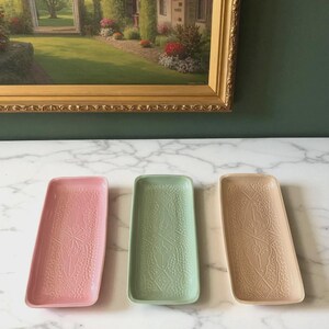 Op de afbeelding: Drie rechthoekige keramische trays in roze, groen en beige, elk met een gedetailleerd reliëf bloemmotief. De trays zijn gerangschikt op een marmeren oppervlak, met een ingelijst schilderij op de achtergrond.