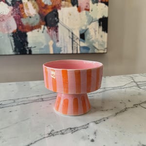 May include: A pink ceramic bowl on a pedestal, decorated with vertical orange stripes. The bowl has a wide, shallow top and a short, flared base. The stripes are evenly spaced. The bowl is likely handmade and suitable for serving or display, with a height of approximately 10 cm.