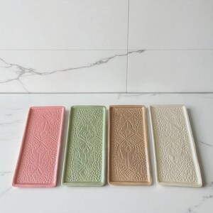 Può includere: Quattro vassoi rettangolari in ceramica rosa, verde, beige e crema. Ogni vassoio presenta un motivo ornato in rilievo. I vassoi sono disposti su una superficie di marmo bianco, offrendo un elemento elegante e decorativo.