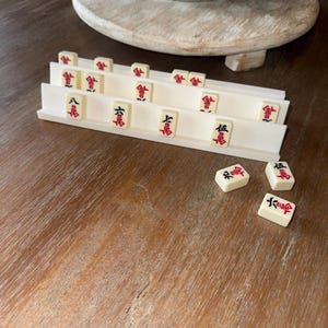 May include: A set of ivory-colored Mahjong tiles with red Chinese characters, displayed in a white tiered holder. Several tiles are scattered on a wooden surface. The game pieces are arranged near a decorative ceramic pot on a wooden stand.