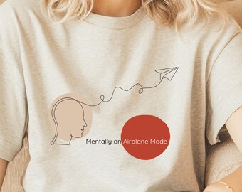 May include: A light gray short-sleeve t-shirt featuring a black line art graphic of a human head profile connected by a wavy line to a paper airplane. A solid red circle is positioned next to the text Mentally on Airplane Mode.