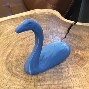 K&ouml;nnte beinhalten: Eine blaue 3D-gedruckte Skulptur einer mythischen Kreatur, die einem langhalsigen Dinosaurier &auml;hnelt, auf einer Holzoberfl&auml;che.