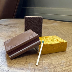 May include: Three small decorative boxes with intricate designs. The boxes are brown, gold, and a darker brown. A matchstick is laying on the table next to the boxes.