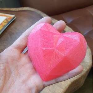 Puede incluir: Un objeto en forma de coraz&oacute;n rosa brillante y facetado. El coraz&oacute;n tiene un dise&ntilde;o geom&eacute;trico con m&uacute;ltiples superficies planas. El objeto se sostiene en una mano. El fondo incluye una mesa de madera y un sof&aacute; de cuero.