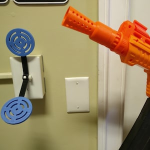 May include: An orange and black toy gun with a large magazine is aimed at a blue target attached to a light switch. The target is made of two blue circular pieces connected by a black strap.