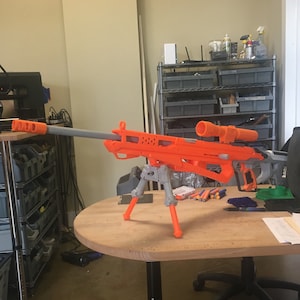 May include: An orange and gray toy sniper rifle with a bipod stand. The rifle has a scope and a long barrel.