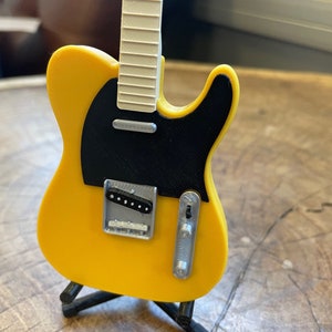 May include: A yellow electric guitar with a black pickguard and a white fretboard. The guitar is on a black stand.