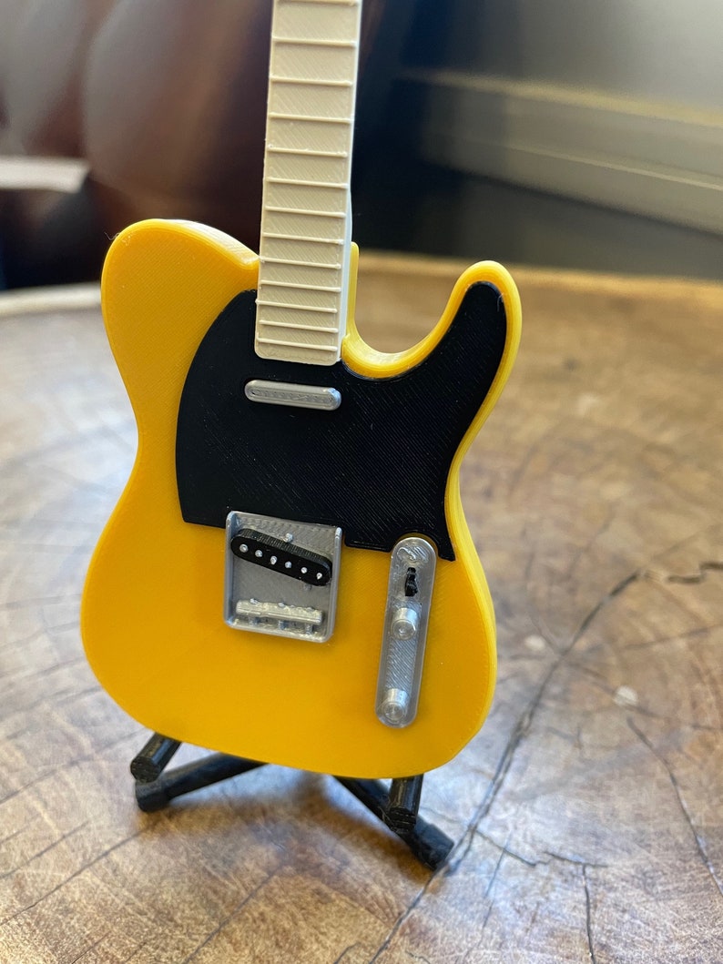 May include: A yellow electric guitar with a black pickguard and a white fretboard. The guitar is on a black stand.