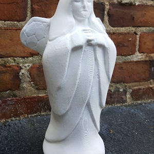 May include: A white angel statue with folded hands and detailed wings. The sculpture has a flowing robe and a serene expression. The statue is set against a brick wall background.
