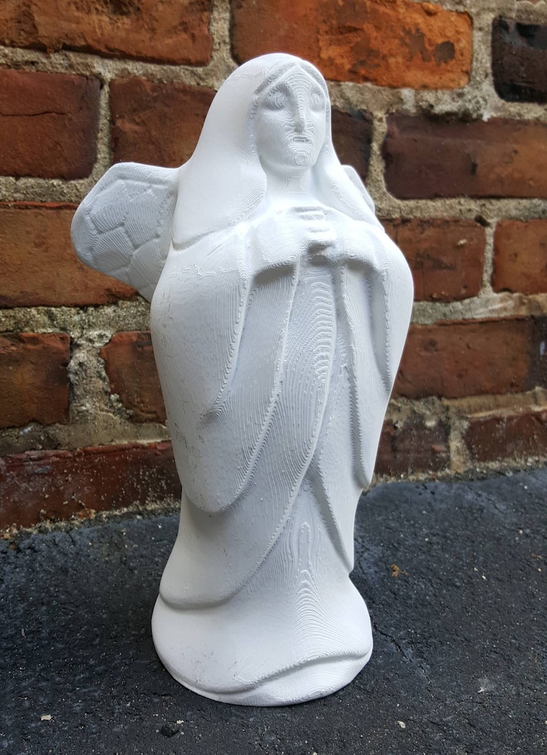 May include: A white angel statue with folded hands and detailed wings. The sculpture has a flowing robe and a serene expression. The statue is set against a brick wall background.