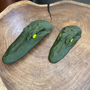 May include: Two green alligator head sculptures with yellow eyes. The sculptures are made of a textured material and are sitting on a wooden surface.