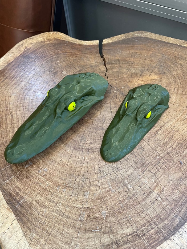 May include: Two green alligator head sculptures with yellow eyes. The sculptures are made of a textured material and are sitting on a wooden surface.