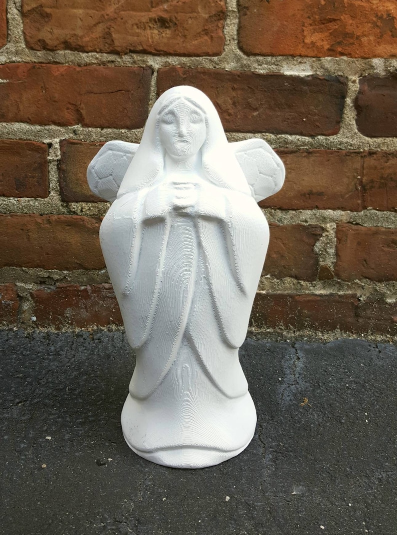 May include: A white angel statue with folded hands and detailed wings. The sculpture has a flowing robe and is set against a brick wall background. The angel is approximately 10 inches tall.
