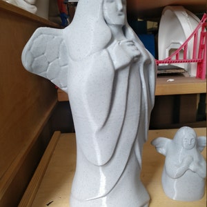 May include: A light gray angel statue with detailed wings and flowing robes. The statue is standing on a wooden surface. A smaller angel statue is also present. The statues have a speckled texture.