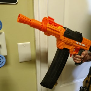 May include: An orange and black toy blaster gun with a large black magazine. The toy gun is held by a person. A blue and white target is mounted on the wall.