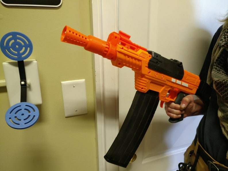 May include: An orange and black toy blaster gun with a large black magazine. The toy gun is held by a person. A blue and white target is mounted on the wall.