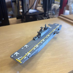 May include: A grey model aircraft carrier with a yellow runway stripe and white markings. The model has detailed structures and is set on a wooden surface. The design suggests a military or hobbyist theme.