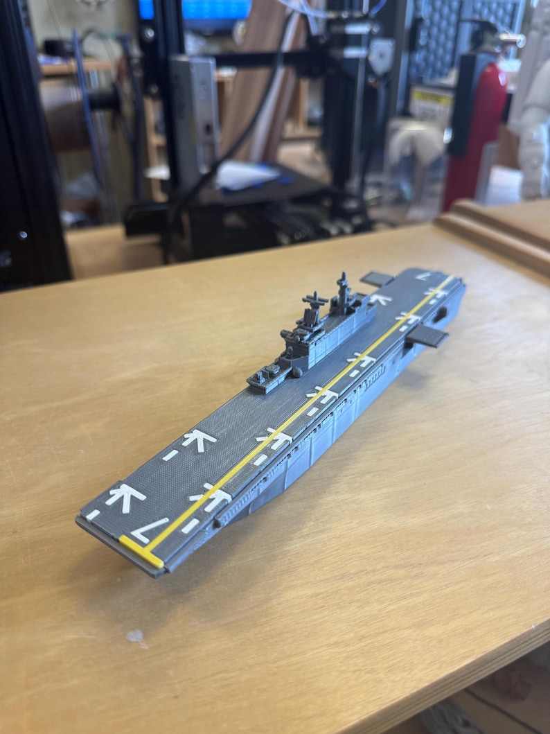 May include: A grey model aircraft carrier with a yellow runway stripe and white markings. The model has detailed structures and is set on a wooden surface. The design suggests a military or hobbyist theme.