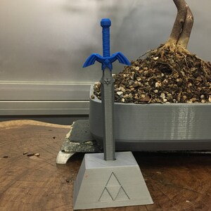 May include: A blue plastic sword with a silver blade and a triangular base with the Triforce symbol. The sword is standing on a wooden surface.