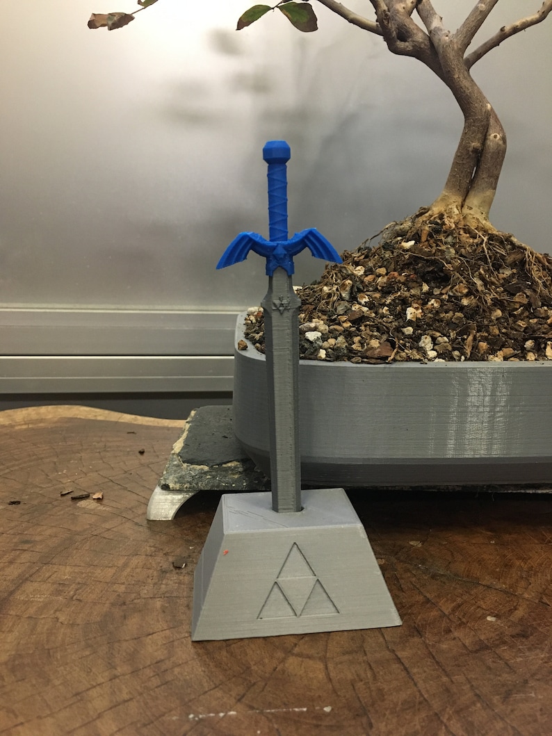 May include: A blue plastic sword with a silver blade and a triangular base with the Triforce symbol. The sword is standing on a wooden surface.