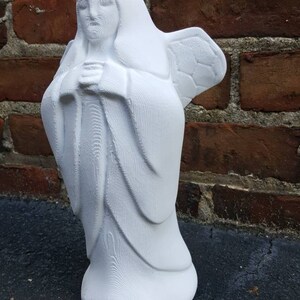 May include: A white angel statue with folded hands and detailed wings. The figure is wearing a long robe and a hood. The statue is made of a solid material and stands on a flat base. The background is a brick wall.