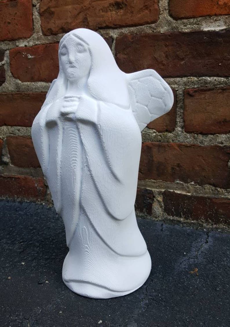 May include: A white angel statue with folded hands and detailed wings. The figure is wearing a long robe and a hood. The statue is made of a solid material and stands on a flat base. The background is a brick wall.