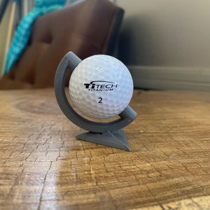 May include: A white golf ball with black text that reads "TiTech Titanium 2" is displayed on a gray, 3D printed globe stand. The stand has a triangular base.