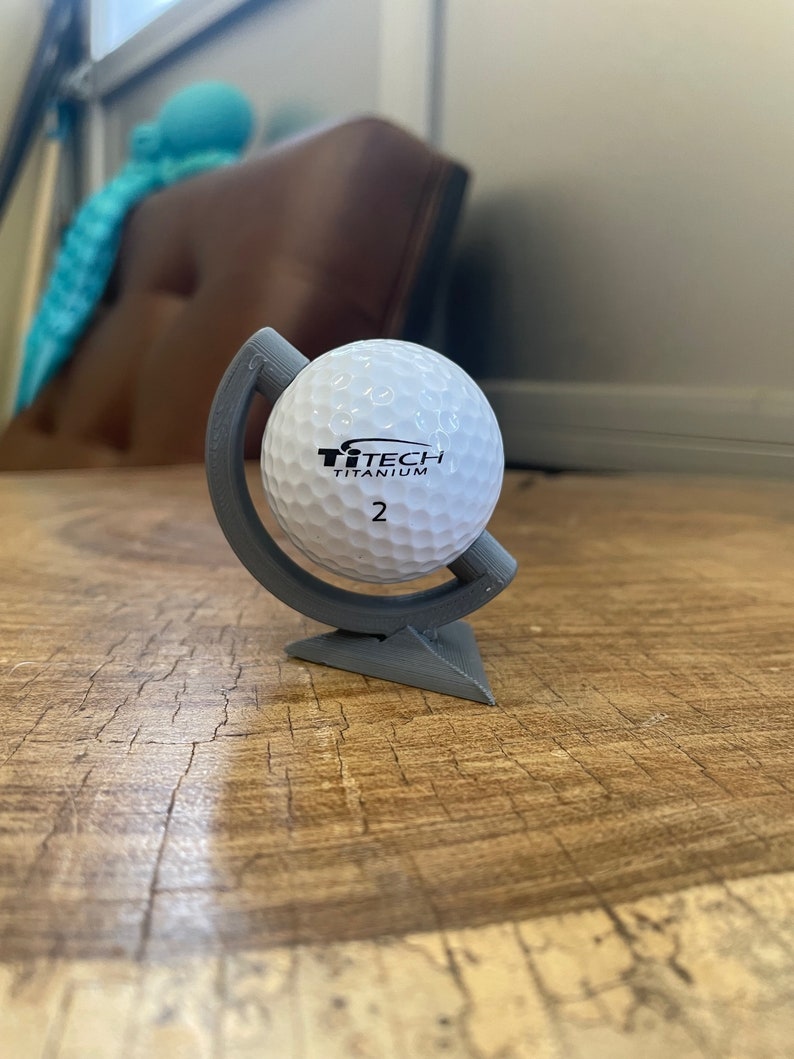 May include: A white golf ball with black text that reads "TiTech Titanium 2" is displayed on a gray, 3D printed globe stand. The stand has a triangular base.