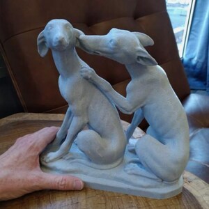 May include: A gray ceramic sculpture of two dogs, one with its head resting on the other's shoulder. The dogs are in a sitting position and appear to be affectionate.
