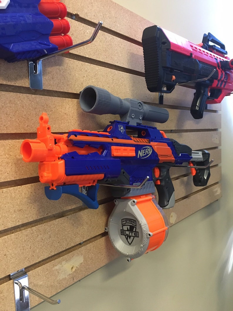 Może przedstawiać: Niebiesko-pomarańczowy blaster Nerf N-Strike Elite z dużym pomarańczowym magazynkiem. Blaster ma celownik teleskopowy i jest zamontowany na drewnianej ścianie z haczykami.