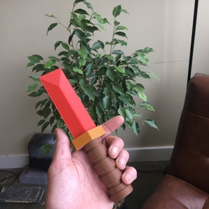 May include: A red and brown plastic toy sword with a textured handle. The sword is held in a person's hand.